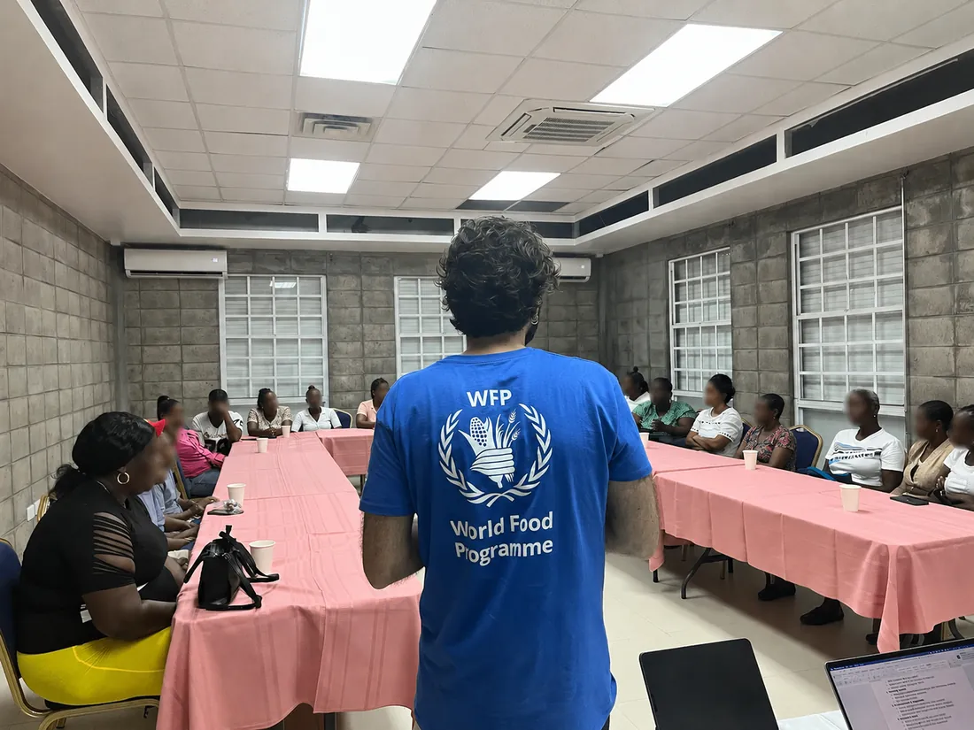 Francesco running a focus group in Grenada, seen from behind in a WFP shirt, with people receiving government assistance seated around tables